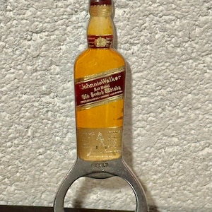 May include: A bottle opener shaped like a Johnnie Walker whisky bottle. The opener has a silver metal base with a bottle-shaped top featuring a yellow and red label. The label reads "Johnnie Walker" and "Old Scotch Whisky."