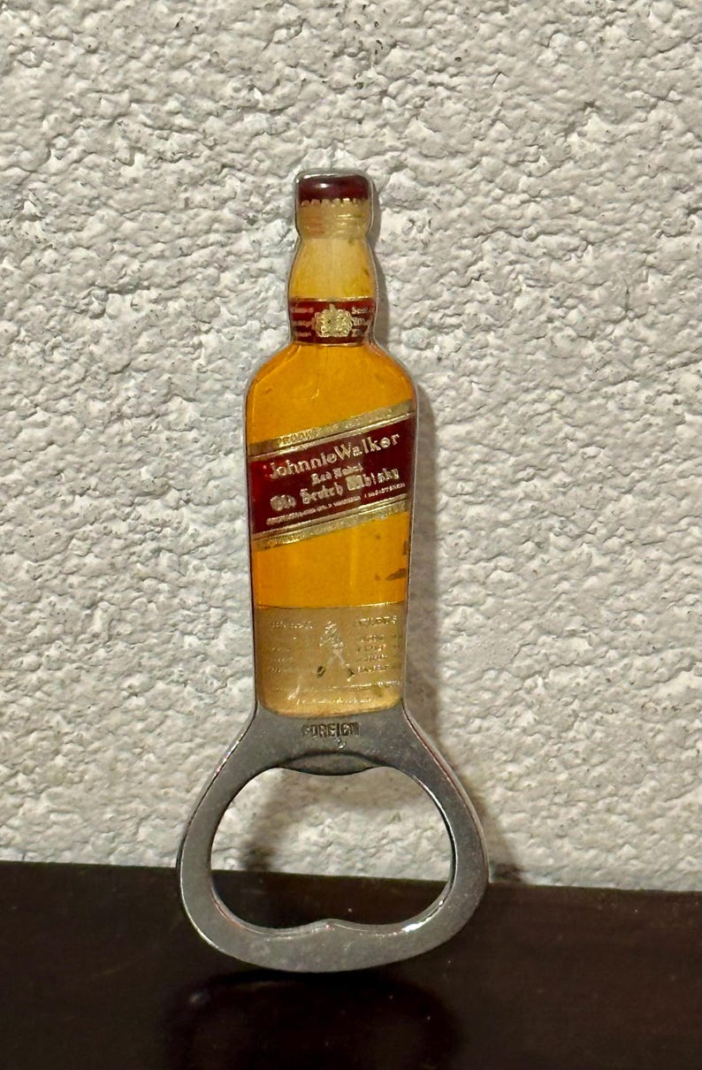May include: A bottle opener shaped like a Johnnie Walker whisky bottle. The opener has a silver metal base with a bottle-shaped top featuring a yellow and red label. The label reads "Johnnie Walker" and "Old Scotch Whisky."