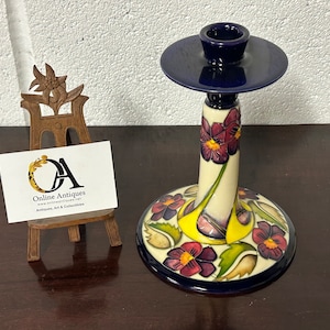 May include: A blue and white ceramic candlestick with a floral design. The candlestick has a round base and a tall, slender stem. The floral design features purple and yellow flowers with green leaves.