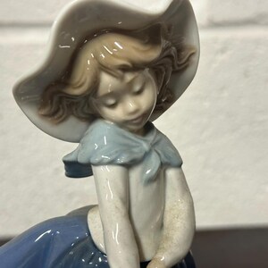 May include: A porcelain figurine of a girl wearing a white hat and a blue dress. She has brown hair and blue eyes.