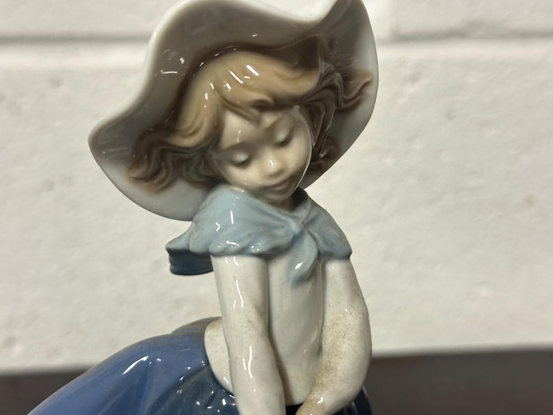 May include: A porcelain figurine of a girl wearing a white hat and a blue dress. She has brown hair and blue eyes.