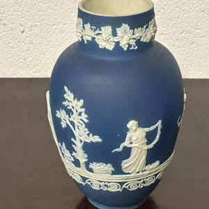 Puede incluir: Un jarrón de cerámica azul y blanco con un diseño en relieve de una mujer y un árbol. El jarrón tiene un borde blanco y una base blanca.