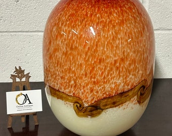 May include: A large, oval-shaped glass vase with a speckled orange and white design. The vase has a brown band around the middle with a swirling pattern.