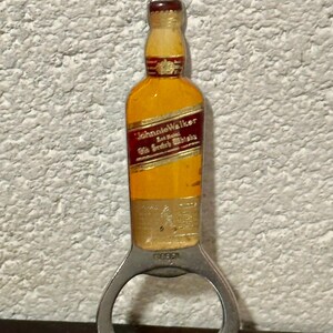 May include: A bottle opener shaped like a miniature Johnnie Walker whiskey bottle. The opener has a silver metal base with a bottle-shaped top in yellow, red, and brown. The text "Johnnie Walker" is visible on the bottle.