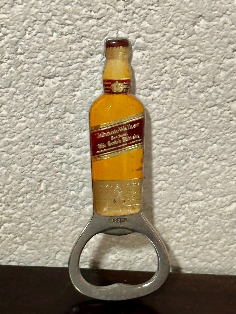 May include: A bottle opener shaped like a miniature Johnnie Walker whiskey bottle. The opener has a silver metal base with a bottle-shaped top in yellow, red, and brown. The text "Johnnie Walker" is visible on the bottle.
