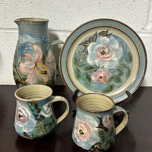 May include: A set of hand-painted pottery featuring a floral design. The set includes a jug, two mugs, and a plate. The pottery is glazed in shades of blue, green, and brown, with pink and white flowers.