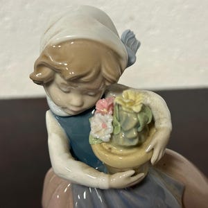 May include: A porcelain figurine of a girl holding a pot of flowers. The girl is wearing a white dress with a blue sash and a white hat. The flowers are pink, yellow, and green.