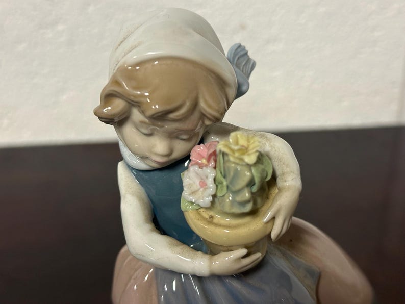 May include: A porcelain figurine of a girl holding a pot of flowers. The girl is wearing a white dress with a blue sash and a white hat. The flowers are pink, yellow, and green.