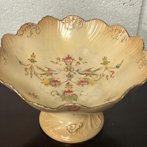 Op de afbeelding: Een crèmekleurige keramische voetkom met een gegolfde rand en gouden rand. De kom is versierd met een bloemenpatroon in tinten roze, groen en blauw.