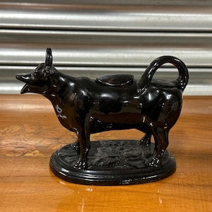 May include: A black ceramic cow creamer with a spout and lid. The cow is standing on a black base with a floral design.