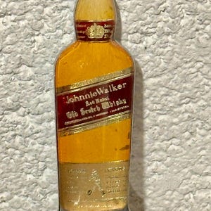 May include: A vintage bottle opener shaped like a Johnnie Walker Red Label whisky bottle. The opener is yellow with a red label and gold lettering. The bottom of the opener has a silver bottle cap remover.