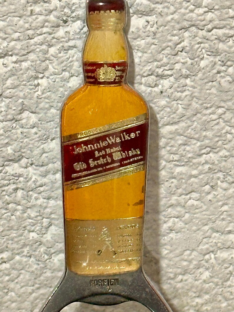 May include: A vintage bottle opener shaped like a Johnnie Walker Red Label whisky bottle. The opener is yellow with a red label and gold lettering. The bottom of the opener has a silver bottle cap remover.