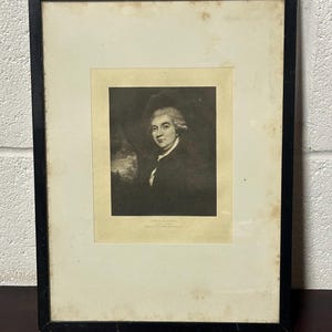 Könnte beinhalten: Gerahmtes Schwarz-Weiß-Porträt eines Mannes in dunklem Mantel und weißer Perücke. Das Porträt ist vor einem hellen Hintergrund platziert und in einem schwarzen Holzrahmen eingefasst. Der Druck ist leicht gealtert.