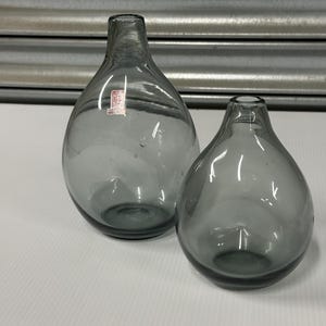 May include: Two gray glass bottles with a rounded body and narrow neck. The bottles are different sizes and sit on a white surface. One bottle has a small red and white label. The background is a gray metal shutter.