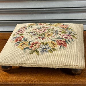 Pode incluir: Um apoio para os pés quadrado com um design floral em ponto de agulha. O tecido bege é adornado com uma coroa de flores rosa, vermelhas, amarelas e azuis. É suportado por quatro pequenos pés de madeira escura.