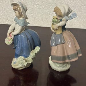 May include: Two porcelain figurines of girls in blue and pink dresses, one holding a basket of flowers and the other holding a potted plant.