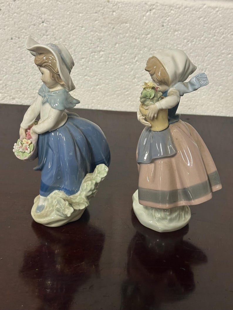 May include: Two porcelain figurines of girls in blue and pink dresses, one holding a basket of flowers and the other holding a potted plant.