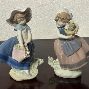 May include: Two porcelain figurines of girls dressed in blue and pink dresses. The girl on the left is holding a basket of flowers and the girl on the right is holding a potted plant.
