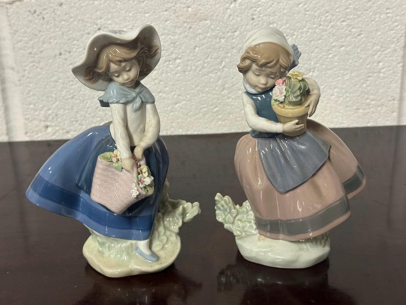 May include: Two porcelain figurines of girls dressed in blue and pink dresses. The girl on the left is holding a basket of flowers and the girl on the right is holding a potted plant.