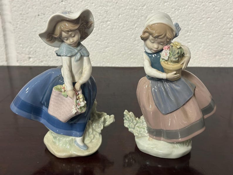 May include: Two porcelain figurines of girls dressed in blue and pink dresses. The girl on the left is holding a basket of flowers and the girl on the right is holding a pot of flowers.