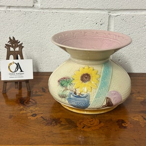 May include: A vintage ceramic vase with a pink rim and a floral design featuring a sunflower, a blue and white patterned pot, and a purple flower. The vase has a woven pattern and is decorated with green leaves.