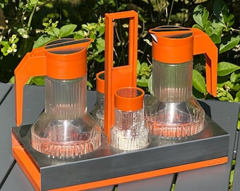 May include: A vintage orange and clear glass condiment set with a metal tray. The set includes two pitchers, two small jars, and a handle. The pitchers and jars have ribbed sides and orange lids. The tray is gray with orange trim.