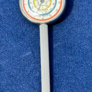 Könnte beinhalten: Ein Kartenmesser aus silberfarbenem Metall mit einem runden Zifferblatt, das mehrere konzentrische Skalen in Orange, Blau und Gelb aufweist. Das Zifferblatt zeigt Texte wie Statute Miles, Nautical Miles und Kilometers. Langer, geriffelter Griff.