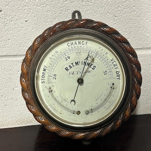May include: A round, wooden framed barometer with a rope-like design around the edge. The barometer face is white with black markings and includes a thermometer. The barometer reads "R&T. MINNES GLASGOW" and has a scale for "STORMY RAIN CHANGE FAIR VERY DRY".