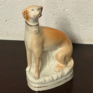 Puede incluir: Figura de perro de cerámica en posición sentada, con una combinación de colores marrón claro y crema. El perro tiene un collar con una etiqueta y está posado sobre una base blanca con ribete dorado. La figura tiene una apariencia detallada y realista.