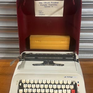 May include: A white Olivetti President portable typewriter with a black keyboard and a red return key. The typewriter is in a burgundy carrying case with a yellow foam insert. The case has a label that says "This Olivetti President Portable Typewriter belongs to" with spaces for a name, address, and telephone number.