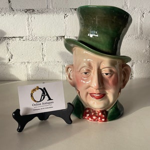 May include: A ceramic figurine of a man's head wearing a green top hat and a red bow tie with white polka dots. The figurine is on a black stand. The text on the card reads "Online Antiques, www.onlineantiques.net, Antiques, Art & Collectibles".