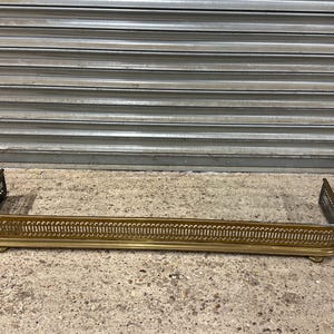 May include: A long, rectangular brass fireplace fender featuring a decorative pierced gallery design. The metal guard has rounded corners and is supported by small, low-profile feet, presenting a protective barrier with a polished metallic finish.