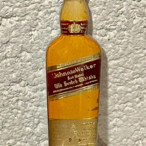 May include: A vintage bottle opener shaped like a Johnnie Walker whiskey bottle. The opener features a brown cap, a yellow bottle with a red label, and a silver-colored opener base. The label reads "Johnnie Walker Old Scotch Whisky."