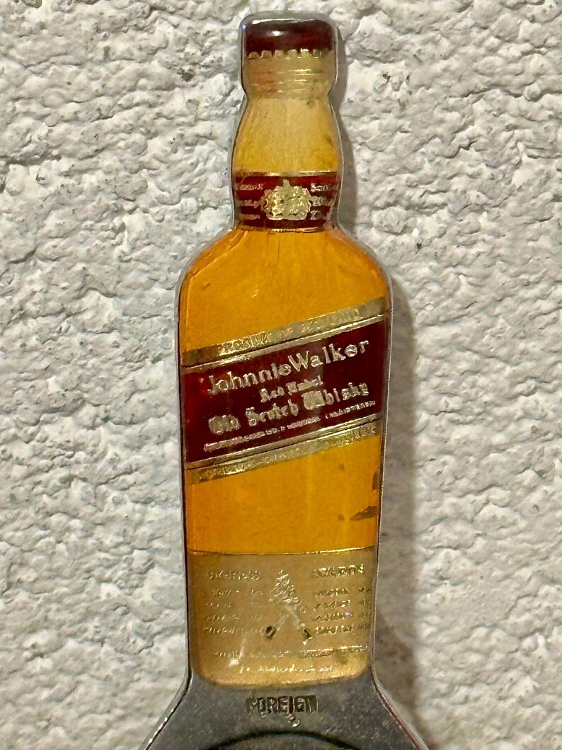 May include: A vintage bottle opener shaped like a Johnnie Walker whiskey bottle. The opener features a brown cap, a yellow bottle with a red label, and a silver-colored opener base. The label reads "Johnnie Walker Old Scotch Whisky."