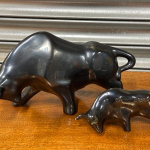 May include: Two dark bronze bull figurines. The larger bull is in a charging stance, while the smaller bull is lowered. Both have curved horns and are on a wooden surface. The sculptures are likely decorative items.