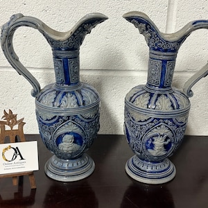 Puede incluir: Dos jarras de cerámica azul y blanca con diseños intrincados. Las jarras tienen un cuerpo bulboso y un cuello alto y delgado. Las asas son curvas y ornamentadas. Las jarras están decoradas con motivos florales y un medallón central.