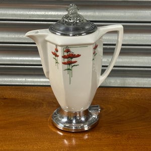 Op de afbeelding: Een witte keramische koffiepot met een zilveren deksel en voet. De pot heeft een bloemenontwerp met rode bloemen en groene stelen. De handgreep is wit en de tuit is schuin voor het schenken.