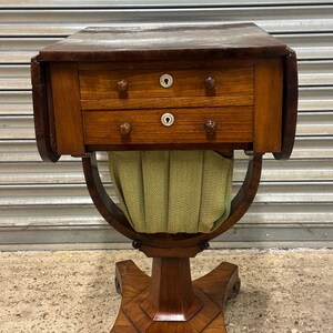 May include: A wooden sewing table featuring a dark brown rectangular top with drop-leaf sides, two stacked drawers with round wooden knobs and white keyhole escutcheons, and a gathered light green fabric basket suspended from a curved frame.