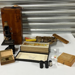 May include: An antique microscope with a wooden case, slides, and accessories. The microscope is black and gold, with a wooden base. Other items include a wooden box, small glass vials, and a slide holder.