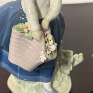 May include: A porcelain figurine of a girl in a blue dress holding a basket of flowers. The girl is smiling and has a pink and white basket with flowers in it. The figurine is on a white base.