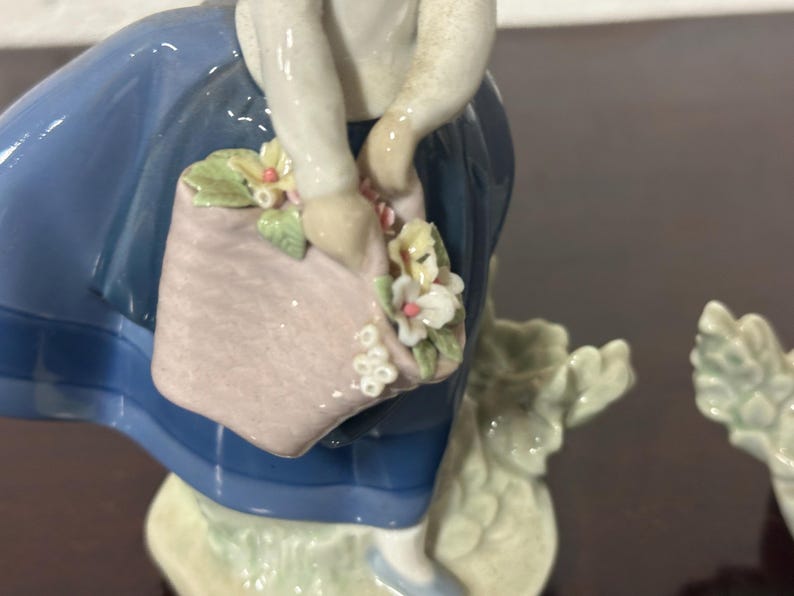 May include: A porcelain figurine of a girl in a blue dress holding a basket of flowers. The girl is smiling and has a pink and white basket with flowers in it. The figurine is on a white base.
