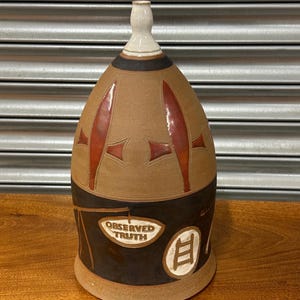 May include: A handcrafted ceramic bottle in brown, tan, and black. It has a white neck, red accents, and the text "OBSERVED TRUTH" on a label. A small ladder design is also present on the bottle.
