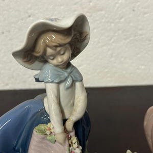 May include: Two porcelain figurines of girls wearing blue dresses and hats. One girl is holding a basket of flowers. The figurines are white with painted details.