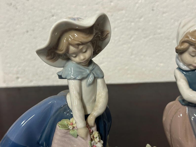 May include: Two porcelain figurines of girls wearing blue dresses and hats. One girl is holding a basket of flowers. The figurines are white with painted details.