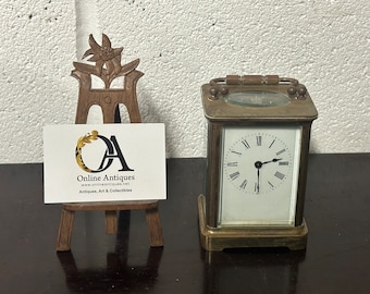 Könnte beinhalten: Eine Messing-Kaminuhr mit weißem Zifferblatt und schwarzen Zeigern. Die Uhr hat ein dekoratives Oberteil mit einem Glasfenster und ein kleines Staffelei mit einer Visitenkarte für "Online Antiques".