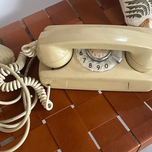 May include: A vintage rotary dial phone with a beige body and a white coiled cord. The phone has a round dial with black numbers 6, 7, 8, 9, and 0.