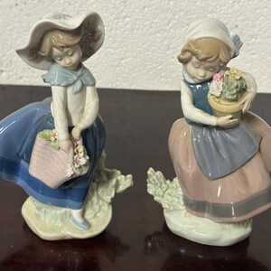 May include: Two porcelain figurines of girls, one wearing a blue and white dress and a hat, holding a basket of flowers, the other wearing a pink and blue dress, holding a pot of flowers.