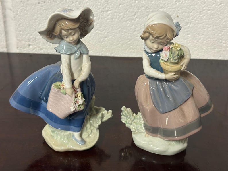 May include: Two porcelain figurines of girls, one wearing a blue and white dress and a hat, holding a basket of flowers, the other wearing a pink and blue dress, holding a pot of flowers.