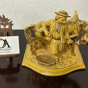 May include: A gold-colored ceramic figurine of a man leading a donkey. The figurine is detailed with a basket and floral accents. The figurine is on a gold-colored base.