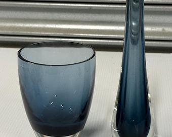 May include: Two pieces of blue glass home decor. One is a short, wide tumbler. The other is a tall, teardrop-shaped vase. Both pieces have a gradient of blue, fading to clear at the base.
